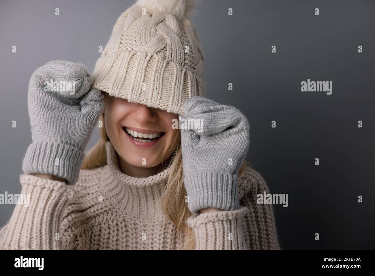 Fröhliche lächelnde Frau versteckt Gesicht unter gestricktem Wollhut. Trendige Wintermode-Kleidung Stockfoto
