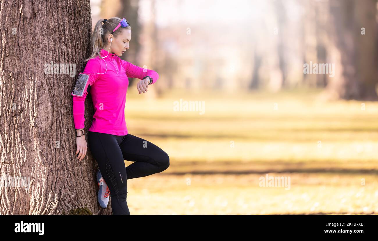Eine junge Sportlerin mit einer athletischen Figur lehnt sich im Park an einen Baum und überprüft beim Laufen ihre Herzfrequenz und hört dabei auch Musik Stockfoto