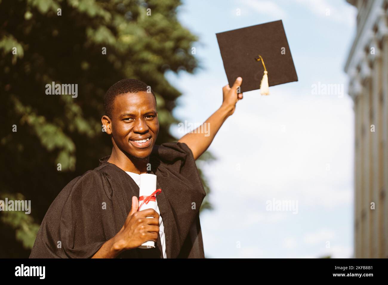 Fröhliche afroamerikanische Absolventen der Universität stehen im Freien in Mantel und in den Händen Hochschulabschluss und schwarzen Hut, verschwommener Himmel Stockfoto