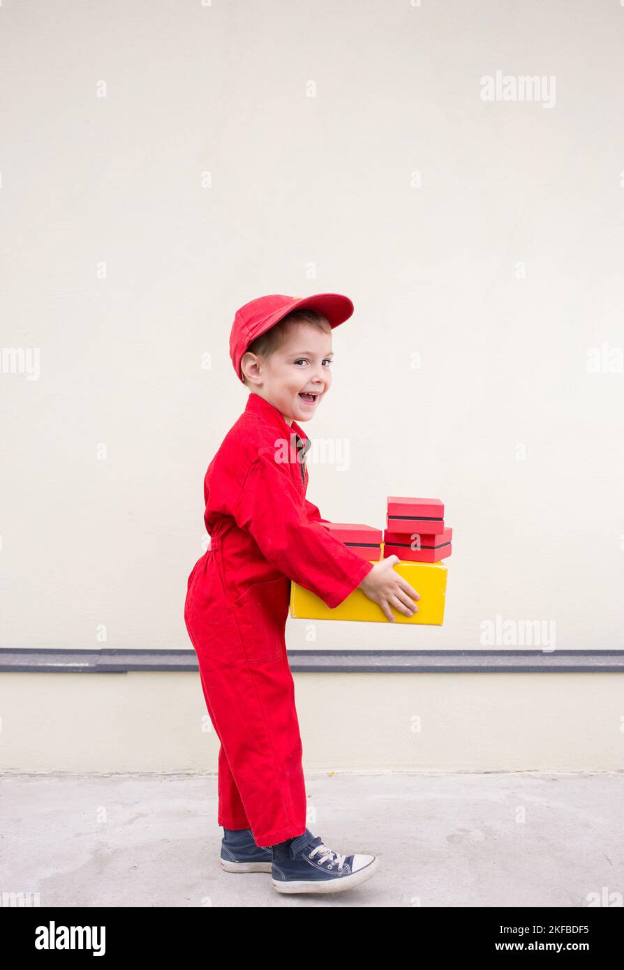 Fröhlicher positiver Junge 3-4 Jahre alt in einer roten Uniform und Mütze mit einem Karton in den Händen. express Lieferung von Glück. Positives Feedback. Spc Stockfoto