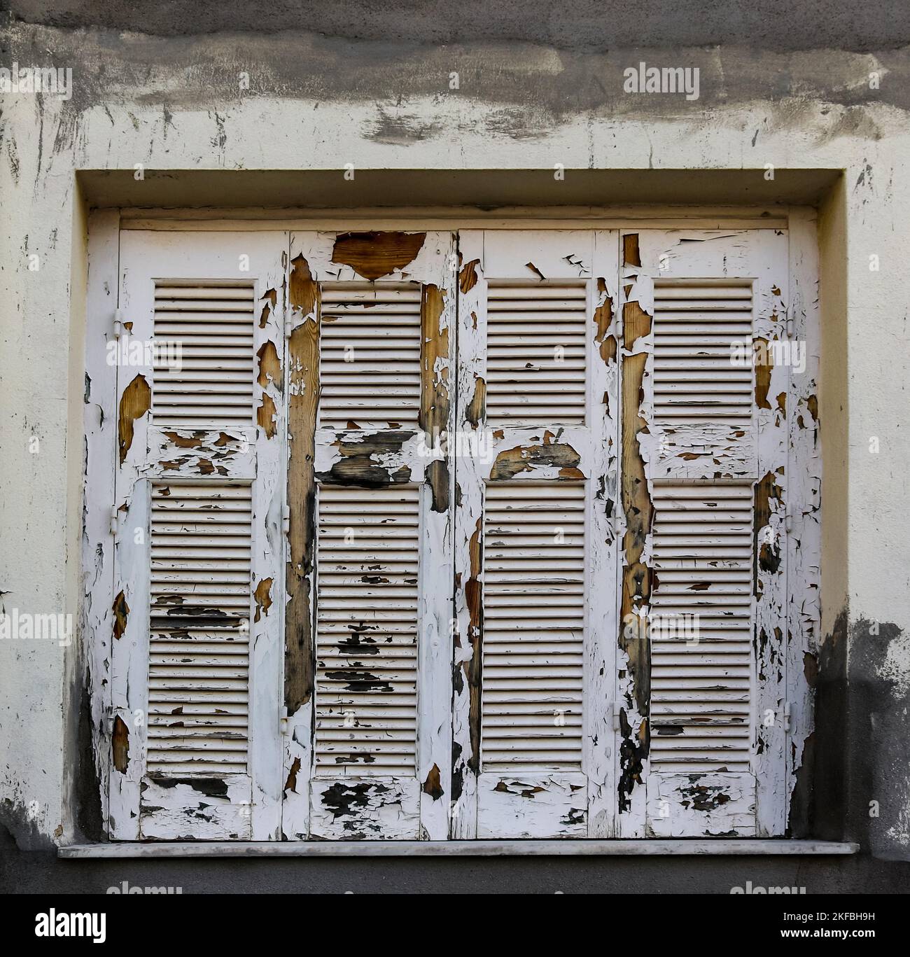 Extrem abgebrochenes und verwittertes weißes Farbwerk auf geschlossenen, grungigen Holzfensterläden in einem Stuckgebäude mit grauem Lack, der reichlich verschmiert ist Stockfoto