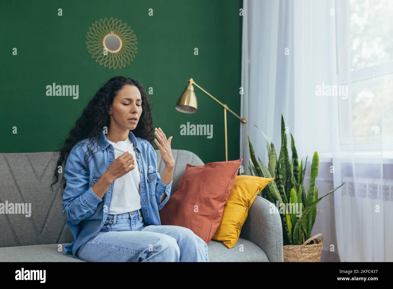 Saisonale Allergie. Eine junge, schöne lateinamerikanische Frau sitzt zu Hause auf der Couch und leidet unter Allergien. Er winkt ihm die Hand ins Gesicht, es ist heiß, es ist schwer zu atmen. Stockfoto
