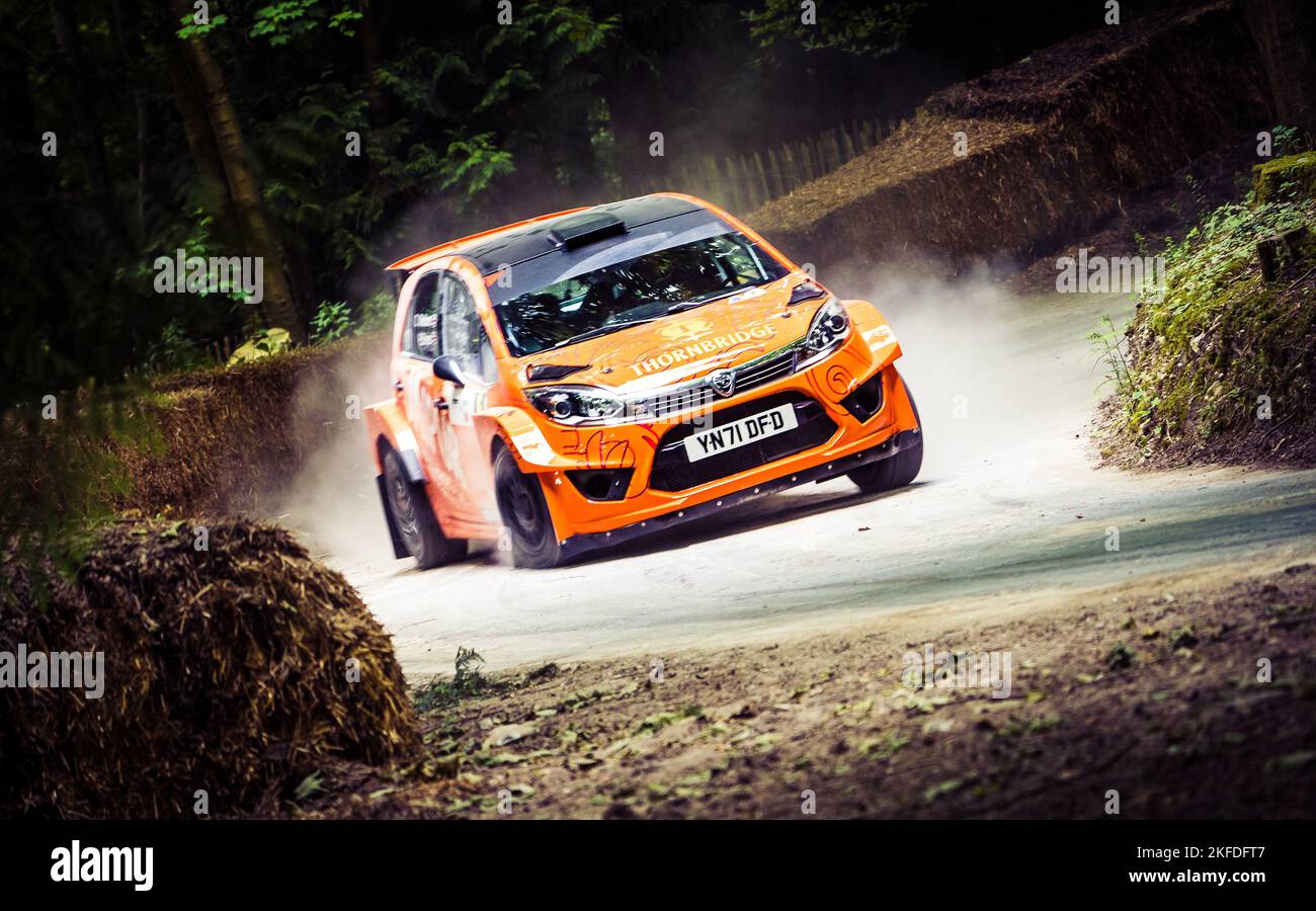 Ein weiß-orangefarbenes Auto, das beim Goodwood Festival of Speed auf der Straße durch einen grünen Wald rast Stockfoto