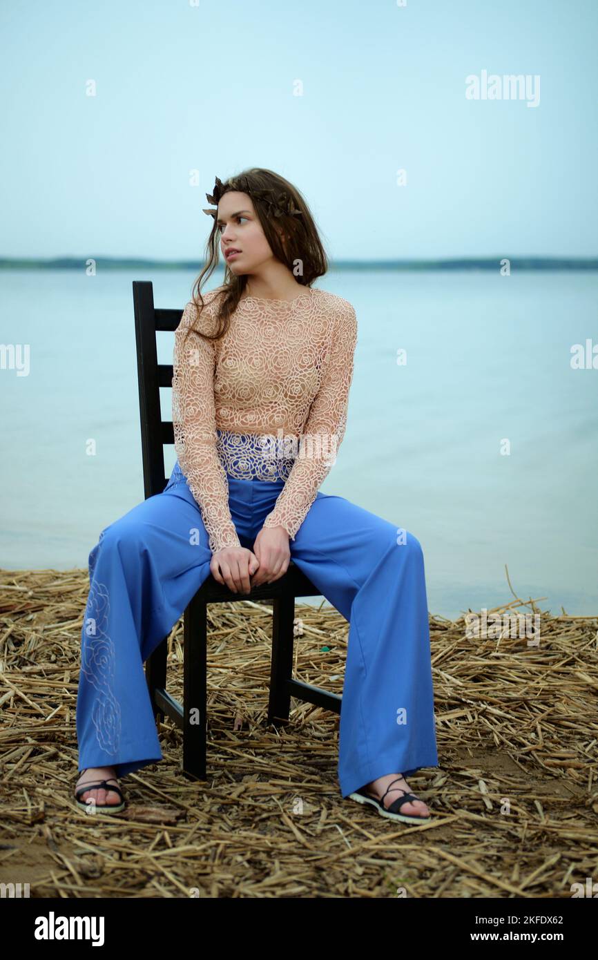 Ruhiges Model in weiten blauen Hosen und Spitzenbluse auf einem Stuhl am Ufer des Sees sitzend Stockfoto