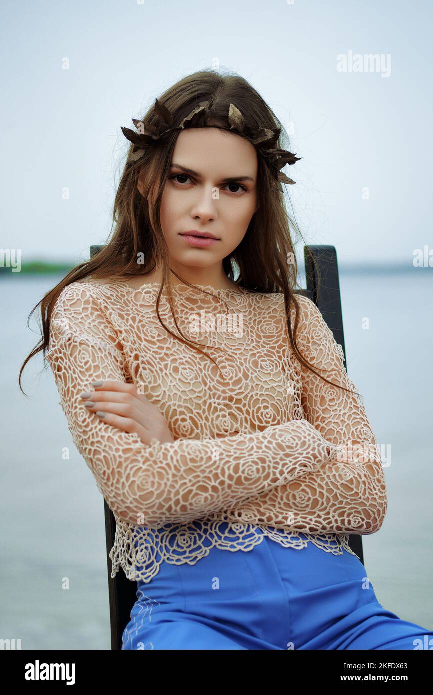Ruhige einsame Frau in weiten blauen Hosen und Spitzenbluse, die an einem bewölkten Tag am Strand auf einem Stuhl sitzt Stockfoto
