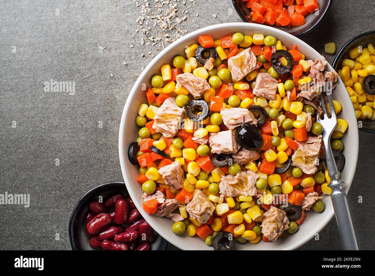 Gesunder gemischter Thunfischsalat mit Hühneraugen, Karotten, Erbsen, Bohnen und Oliven auf grauem Tischhintergrund. Mexikanischer Maissalat. Stockfoto