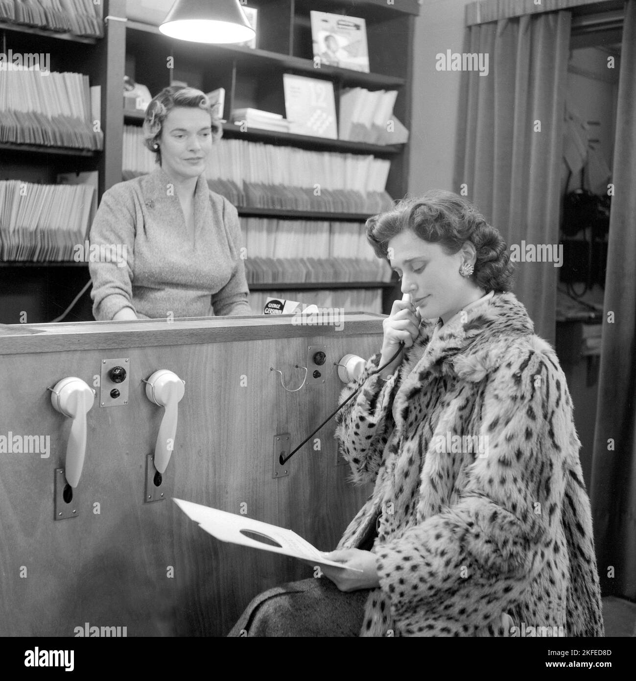 Wie Sie im Jahr 1950s Platten gekauft haben. Die Dame, die ein Telefon hält, hört sich tatsächlich eine Aufzeichnung an. Zu diesem Zeitpunkt war es üblich, dass die Kunden die Aufzeichnungen anhören konnten, bevor sie sich für den Kauf entschieden hatten. Der Plattenladen verfügt über mehrere Hörstationen und eine Frau hinter dem Tresen, die das System verwaltet. Sie hält eine Platte mit dem Namen George Gershwin auf sich. Schweden 1950s. Stockfoto