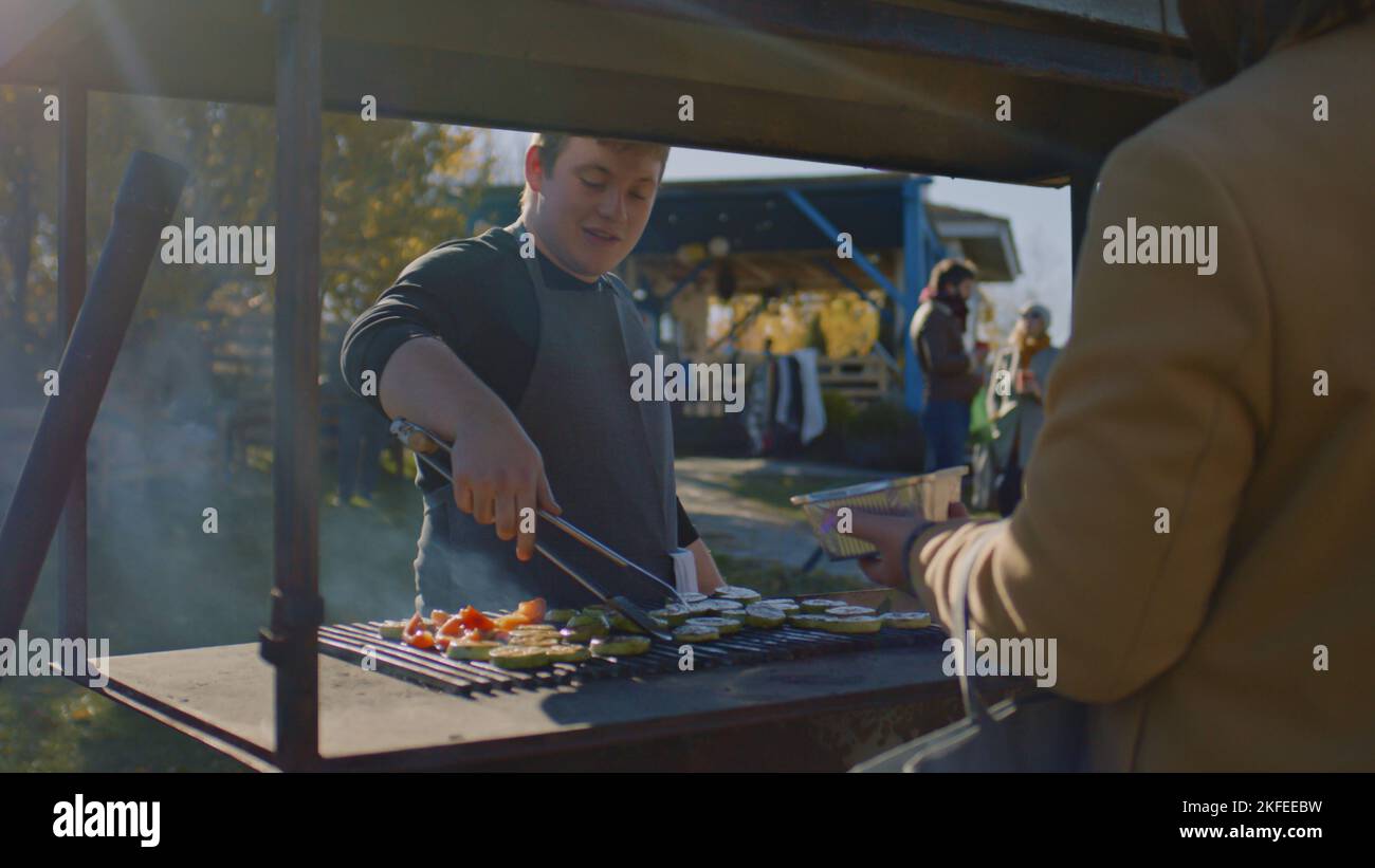 ZAPORIZHYA, UKRAINE - 01. NOVEMBER 2022: Menschen versuchen, Fleisch oder Gemüse auf dem Grill gekocht. Der Erwachsene Mann hebt bereit zum Essen ab und legt eine Portion auf den Teller. Wochenende auf dem lokalen Bauernmarkt. Stockfoto