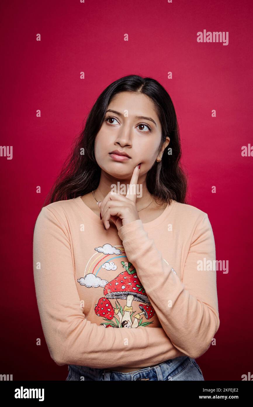 Nachdenkliche junge indische Frau in legerer Kleidung, die an etwas denkt, während sie mit dem Finger auf roten Hintergrund das Kinn berührt. Skeptische Freundin. Stockfoto