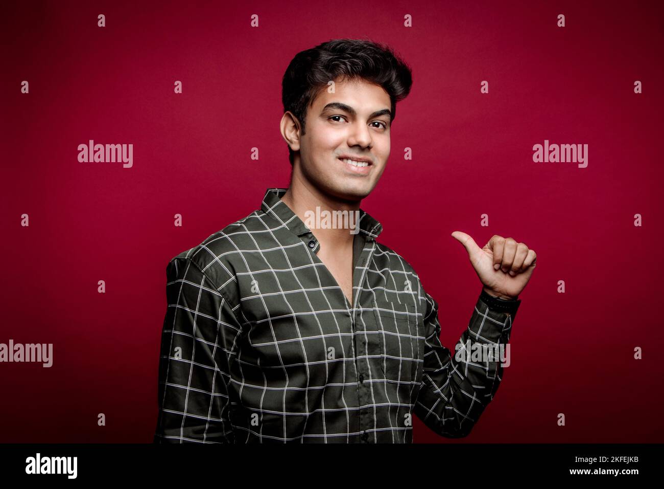 Porträt eines lächelnden jungen indischen Mannes mit kariertem Hemd, der mit dem Daumen auf sich selbst zeigt und vor rotem Hintergrund auf die Kamera blickt Stockfoto