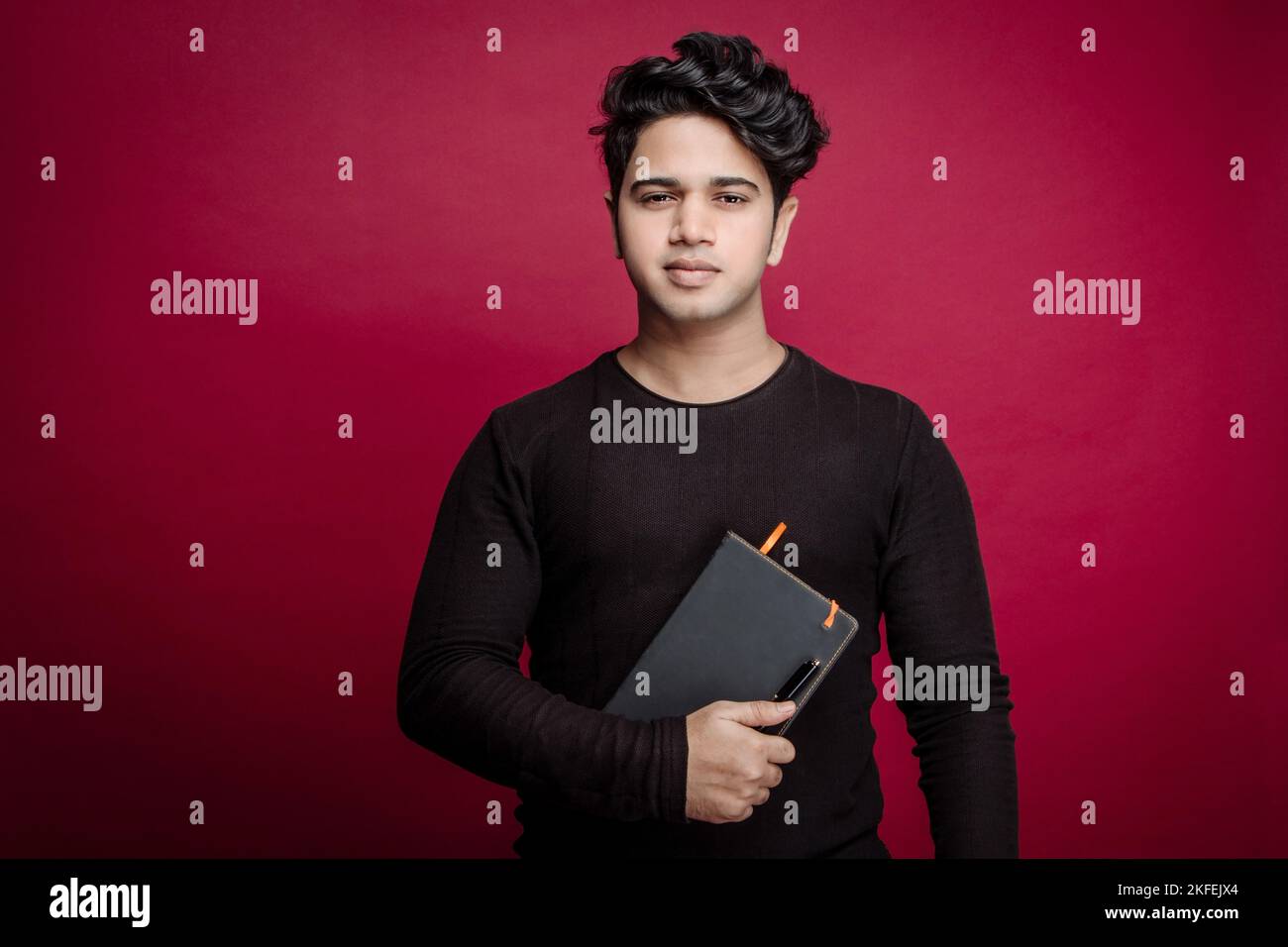 Porträt eines selbstbewussten jungen indischen Geschäftsmannes in einem eleganten, zwanglosen Tagebuch, der vor rotem Hintergrund die Kamera anschaut Stockfoto