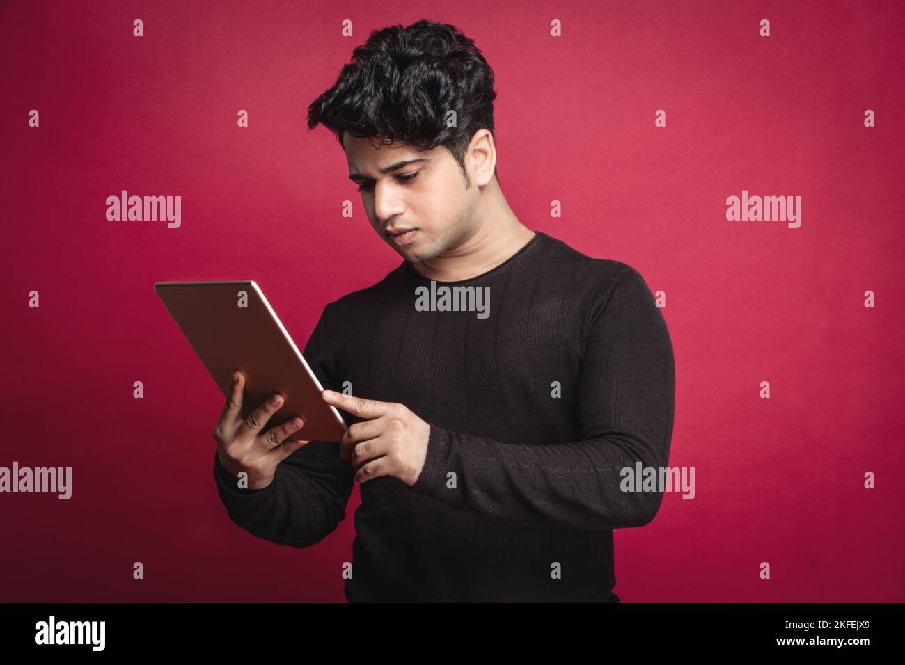 Seriöser junger Inder in einem schwarzen T-Shirt mit einem digitalen Tablet vor rotem Hintergrund Stockfoto