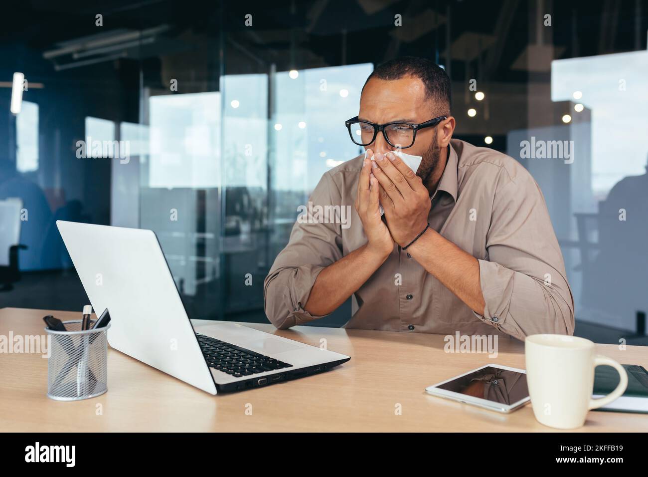 Mann niest und hat eine laufende Nase im Büro bei der Arbeit afroamerikanischer Geschäftsmann krank arbeitet im Büro am Schreibtisch mit Laptop. Stockfoto