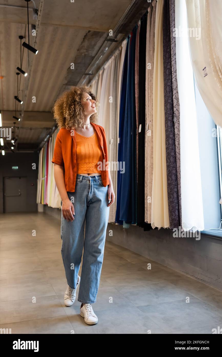 In voller Länge erfreut lockige Frau in Jeans Blick auf bunte Vorhänge im Textilgeschäft Stockfoto