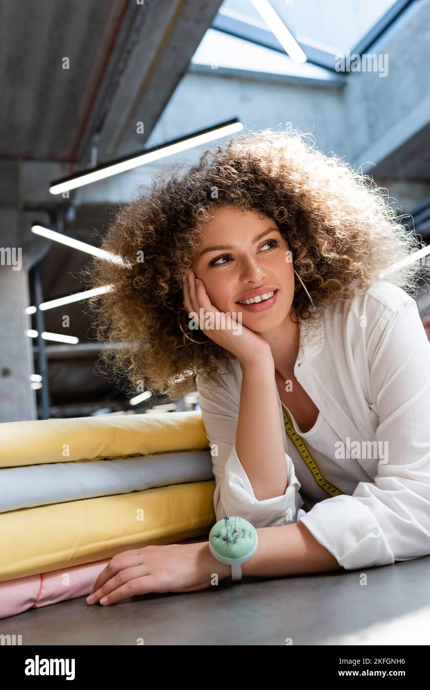 Glückliche Verkäuferin mit Nadelkissen auf der Hand lächelnd in Pastellfarben Stoffrollen im Textilgeschäft Stockfoto
