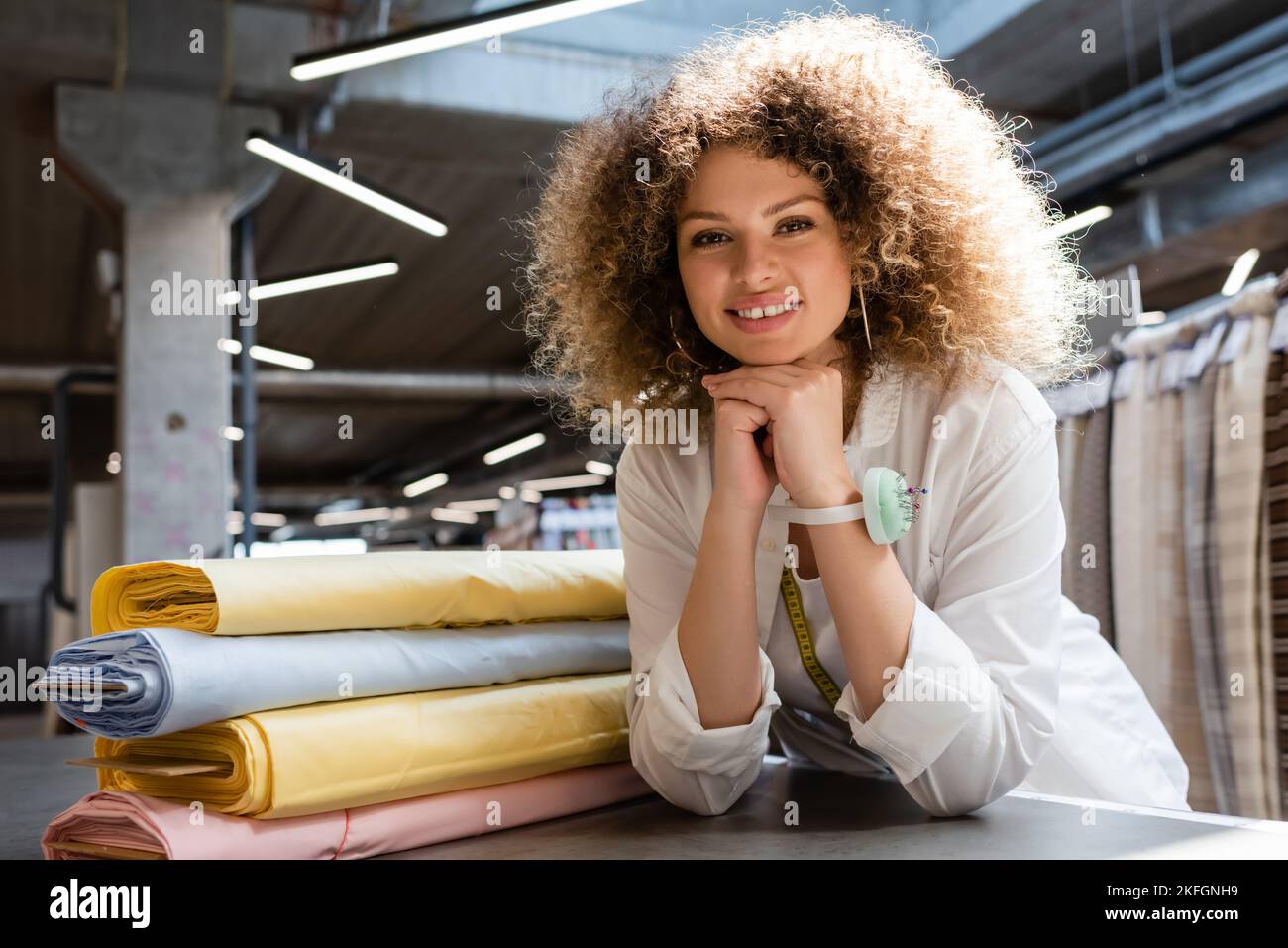Eine erfreuende Verkäuferin mit Nadelkissen auf der Hand, die die Kamera in der Nähe von pastellfarbenen Stoffrollen ansah Stockfoto
