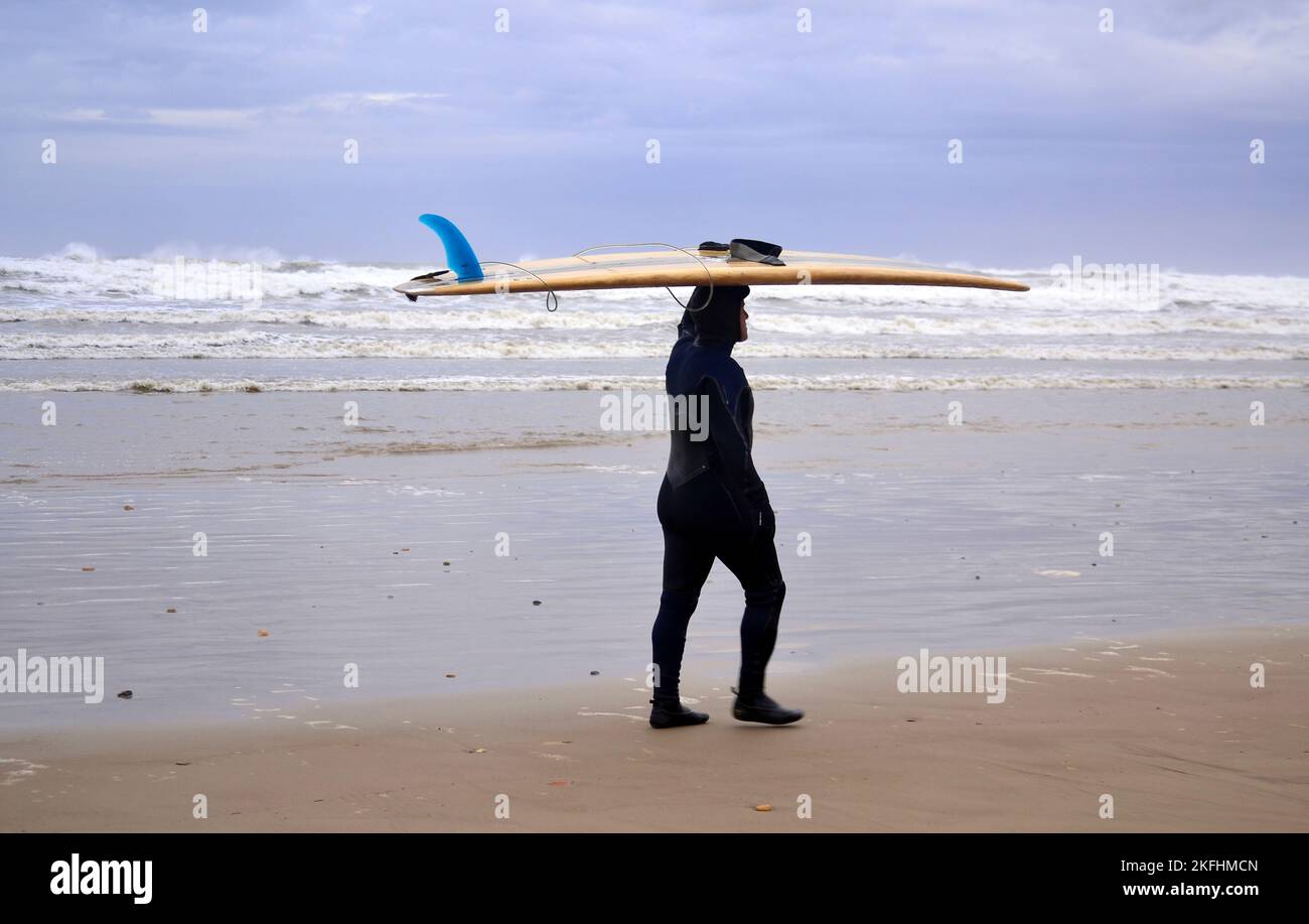 Ein Mann in einem Nassanzug trägt ein Surfbrett auf dem Kopf und läuft am Strand von Saltburn by the Sea, Yorkshire, Großbritannien, entlang. Untersuchungen haben ergeben, dass Reisen in Badeorte der Gesundheit nütze sind. Akademiker von Universitäten befragten Urlauber in Küstenstädten, um nach den Verbindungen zwischen Wohlbefinden und Meer zu fragen. Rund 85 % stimmten ausdrücklich zu oder stimmten darin überein, dass sich das Meer positiv auf Gesundheit und Wohlbefinden auswirkt. Stockfoto