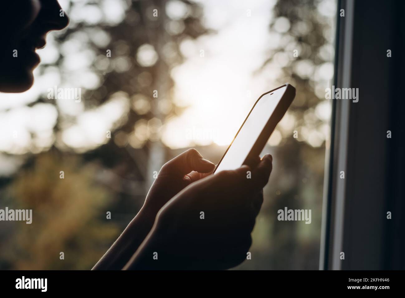 Eine kurze Aufnahme einer jungen Frau, die im Fensterbrett sitzt und das Mobiltelefon hält und die herbstliche Natur während des Sonnenuntergangs genießt. Markt für Smartphone-Anwendungen, Podca Stockfoto