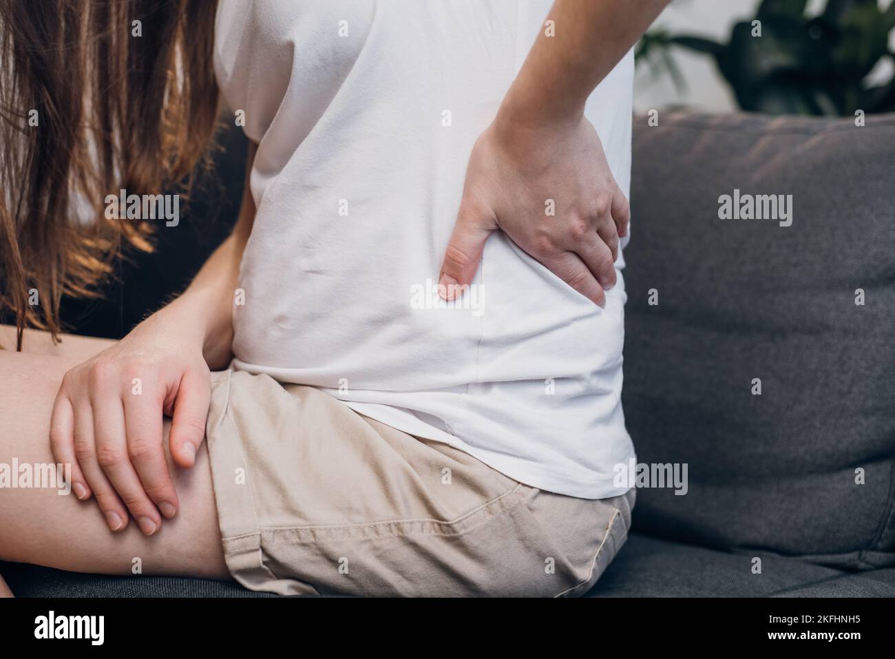 Nahaufnahme einer ungesunden jungen Frau mit Rückenschmerzen, Osteochondrose, die sich mit der Hand zu Hause auf dem grauen Sofa berührt Stockfoto