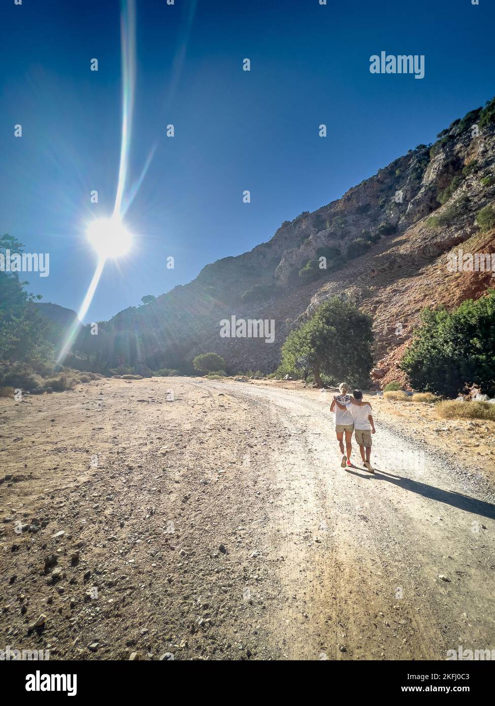 Rückansicht von Mutter und Sohn, die während eines sonnigen Tages auf einer unbefestigten Straße in Richtung Berg gehen Stockfoto