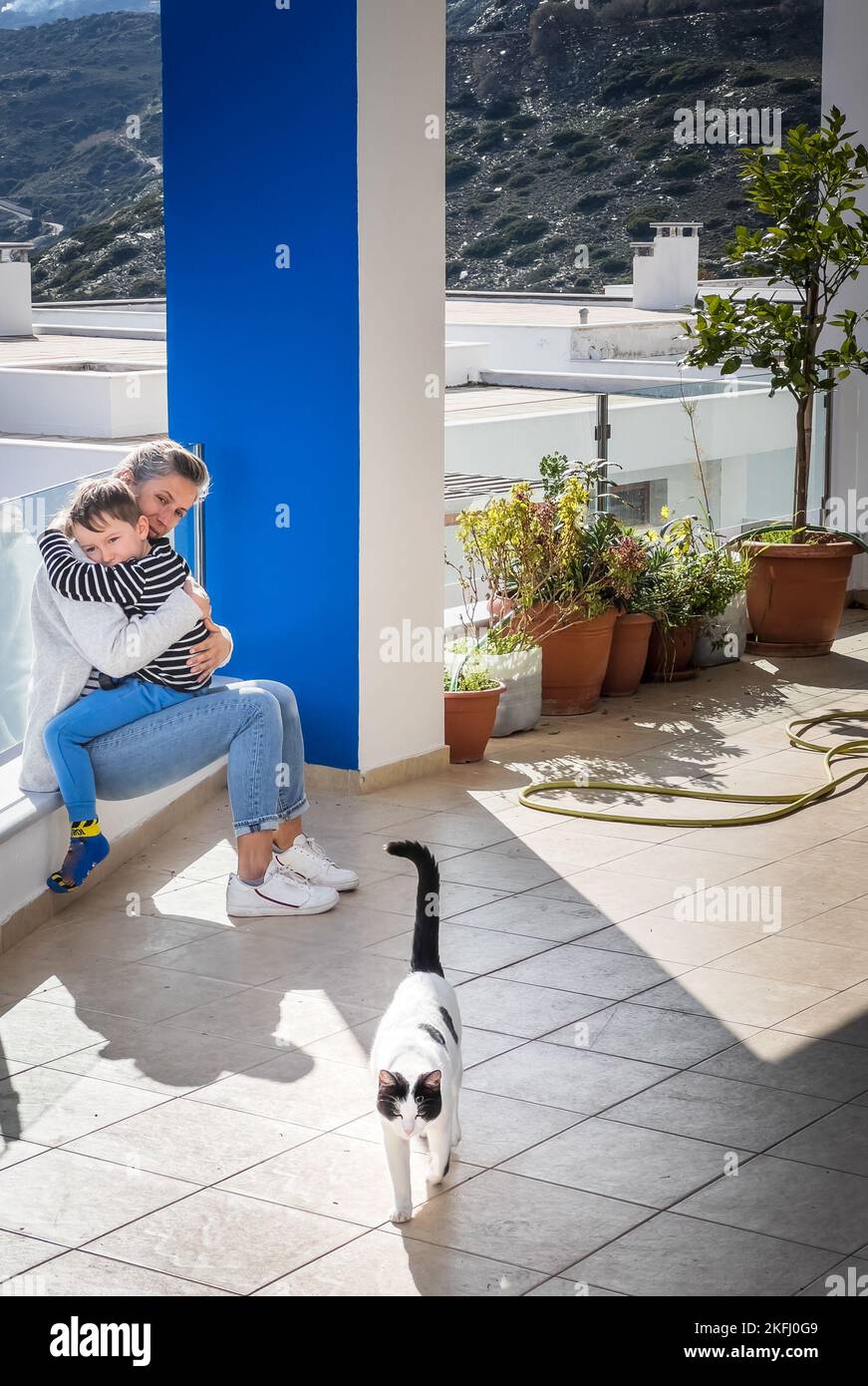 Liebende kaukasische Mutter mittleren Alters umarmt Sohn und Blick auf Katze, während auf dem Balkon sitzen an sonnigen Tagen Stockfoto
