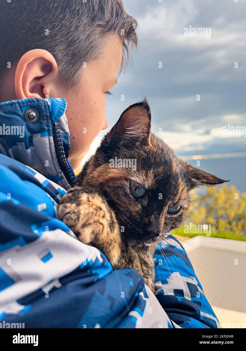 Nahaufnahme eines liebenden Jungen im blauen Mantel, der die Katze im Winter am wolkigen Himmel umarmt Stockfoto