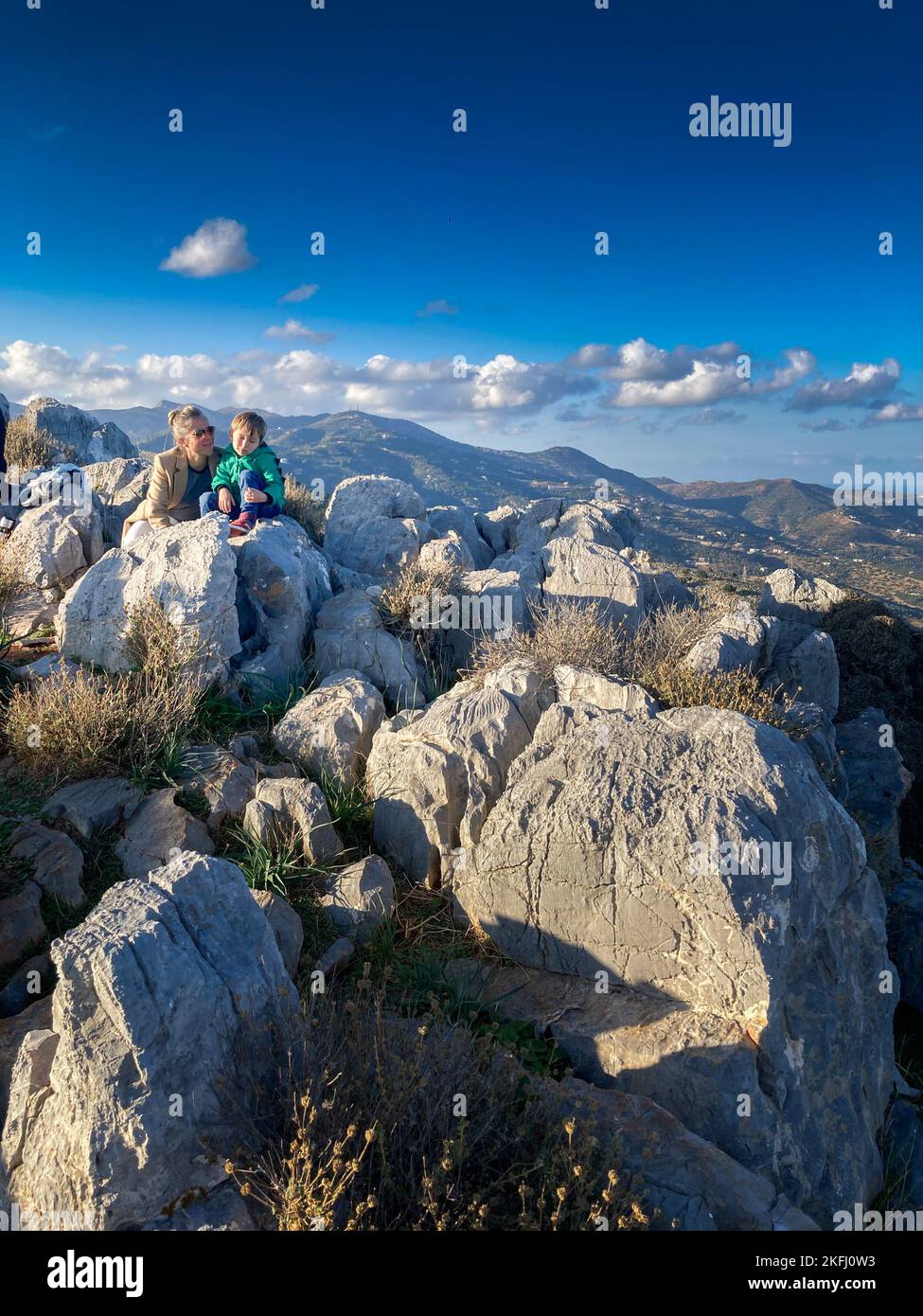 Kaukasische Mutter mit Sohn, die an sonnigen Tagen auf einem felsigen Berg vor blauem Himmel sitzt und den Urlaub genießt Stockfoto