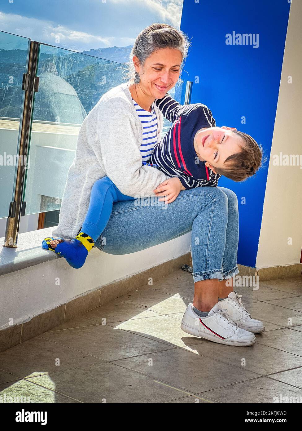Liebende kaukasische Mutter, die auf den niedlichen Sohn schaut, der die Augen schließt und Gesicht macht, während sie auf ihrem Schoß auf dem Balkon sitzt Stockfoto