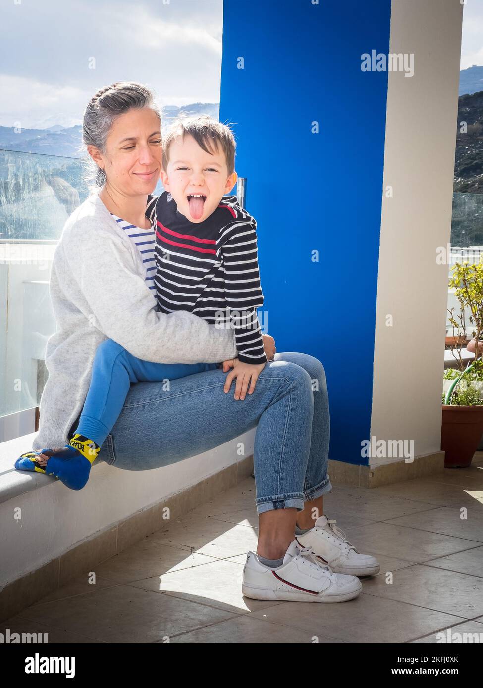 Porträt eines verspielten niedlichen kaukasischen Jungen, der aus der Zunge ragt, während er auf dem Schoß seiner Mutter auf dem Balkon sitzt Stockfoto