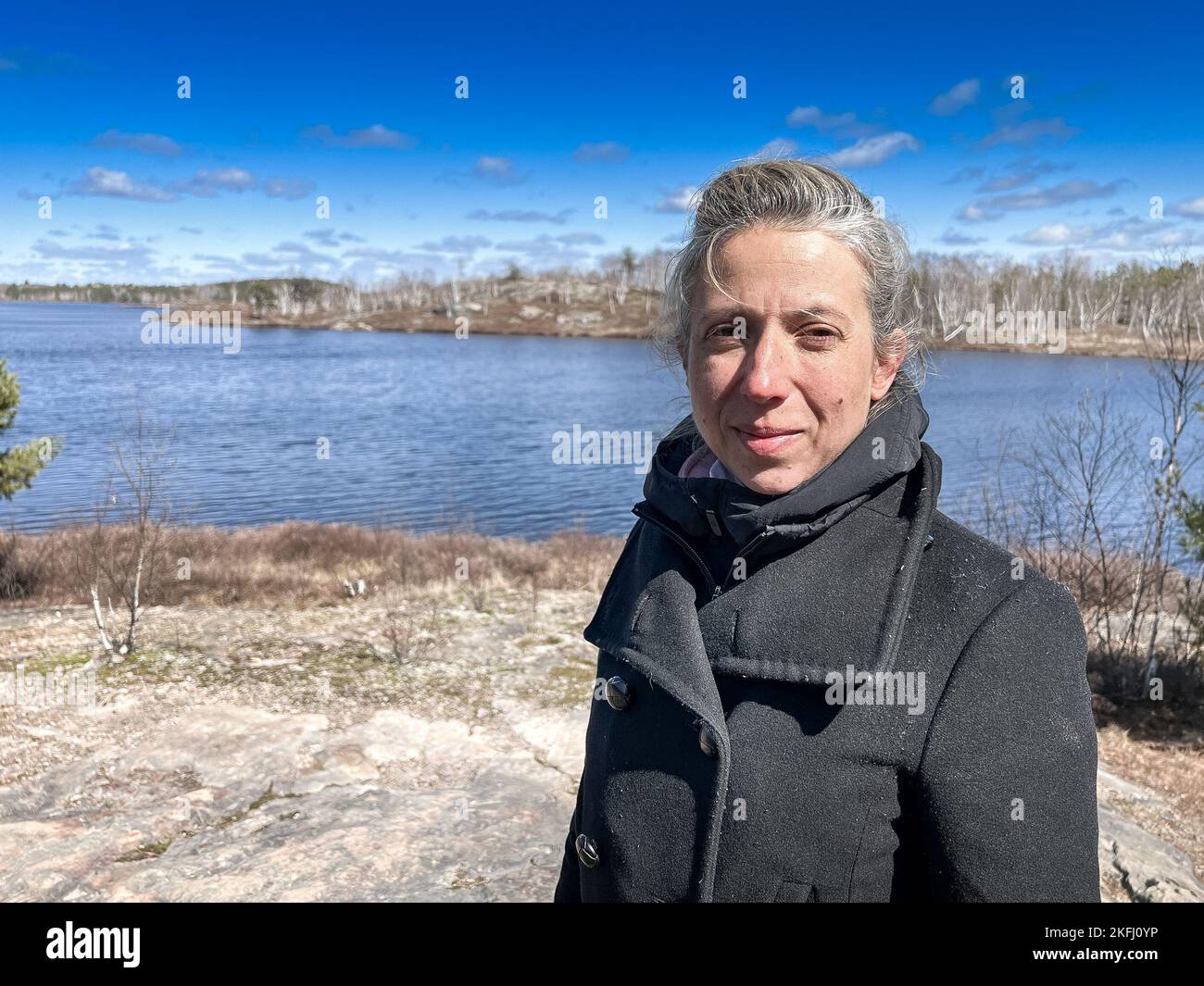 Porträt einer schönen Frau mittleren Erwachsenen in Winterjacke, die an sonnigen Tagen gegen den See und den blauen Himmel steht Stockfoto