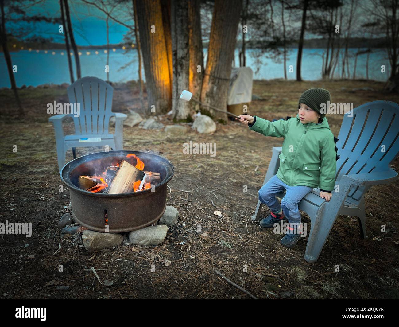 Junge in warmer Kleidung, der während des Sonnenuntergangs im Wald auf einem Adirondack-Stuhl sitzt und über dem Lagerfeuer Sumpflaibe röstet Stockfoto