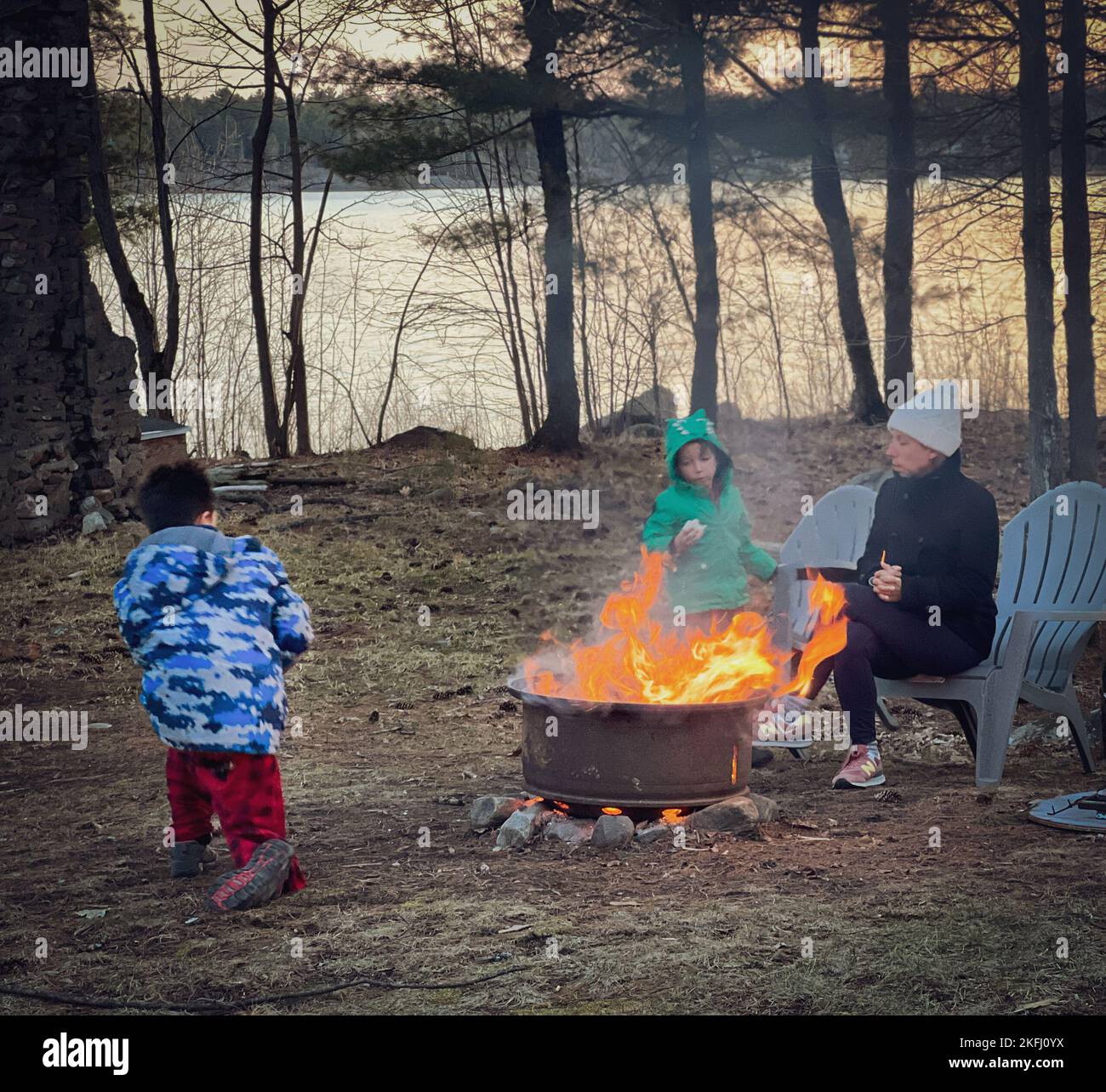 Mutter sitzt auf einem Stuhl und Söhne spielen während des Campens auf dem Land gegen Bäume und See im Wald während des Sonnenuntergangs Stockfoto