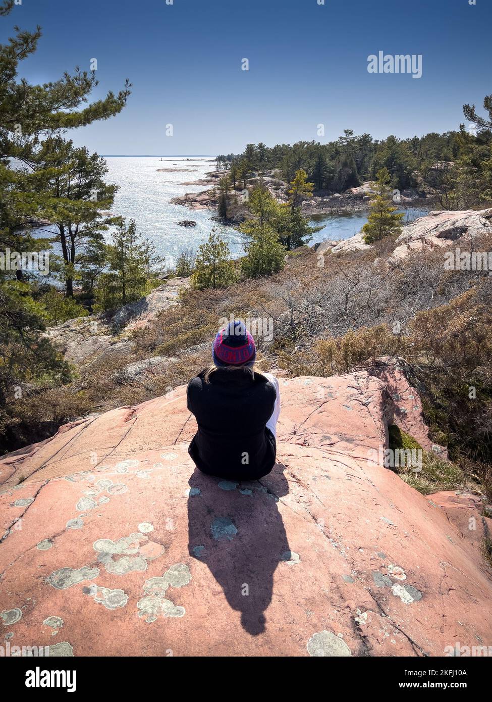 Rückansicht einer Frau mittleren Erwachsenen, die warme Kleidung trägt und das Meer betrachtet, während sie an sonnigen Tagen auf Felsformationen sitzt Stockfoto