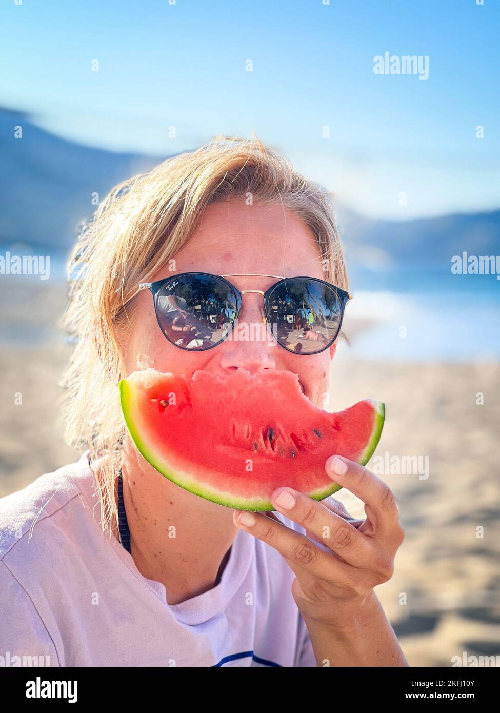 Nahaufnahme eines Porträts einer Frau mittleren Erwachsenen, die eine Sonnenbrille trägt und während der Sommerferien am Strand Wassermelonenscheiben isst Stockfoto
