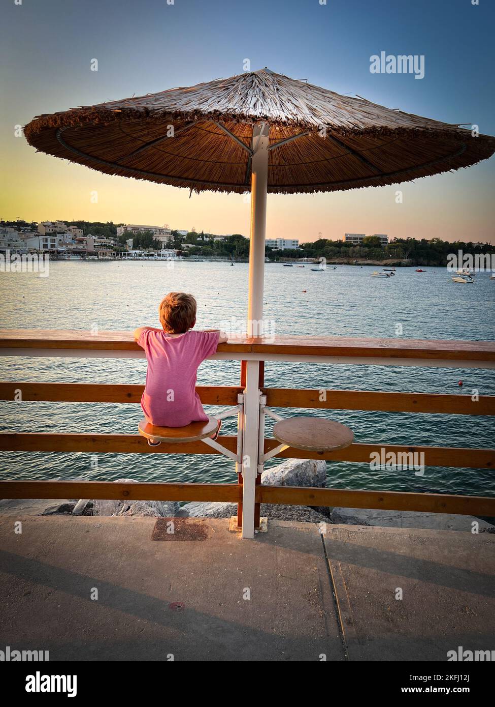 Rückansicht des kaukasischen Jungen, der die Meereslandschaft gegen den Himmel betrachtet, während er während des Sonnenuntergangs am Geländer unter dem Sonnenschirm sitzt Stockfoto