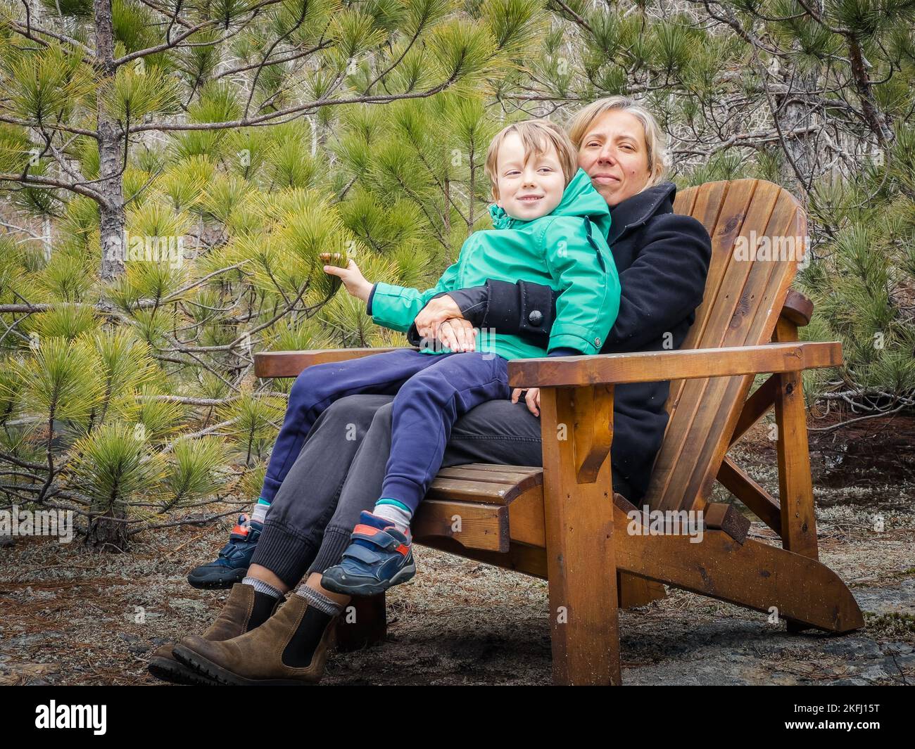 Lächelnde Mutter und Sohn in Wintermänteln sitzen auf einem Holzstuhl gegen üppige Pflanzen, die während des Urlaubs im Wald wachsen Stockfoto