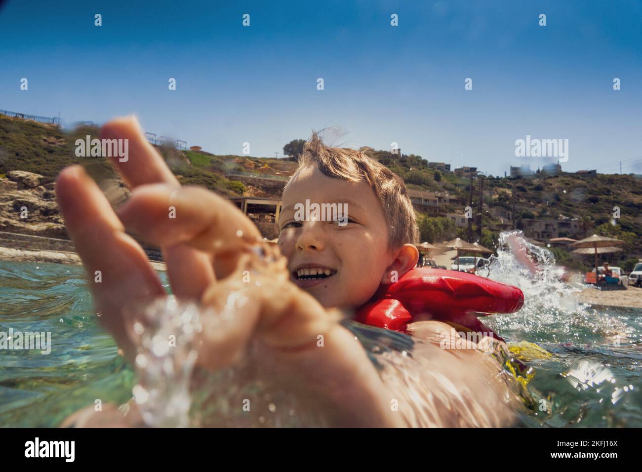 Fröhlicher kaukasischer Junge mit Schwimmweste, der im Meer gegen den Berg und den klaren blauen Himmel während der Sommerferien schwimmt Stockfoto