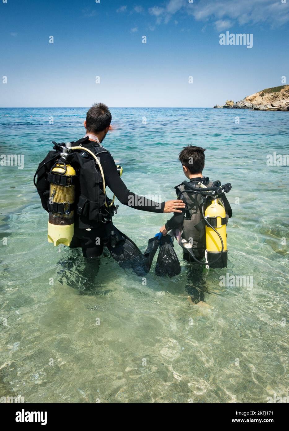 Vater und Sohn tragen Tauchausrüstung und betrachten die Aussicht, während sie an sonnigen Tagen in der Meereslandschaft und gegen den blauen Himmel stehen Stockfoto