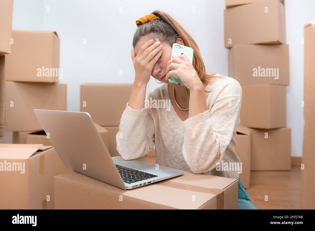 Kaukasische Frau mit Kopfschmerzen beim Umzug in ein neues Zuhause mit Laptop und telefoniert mit einem Berater. Stockfoto
