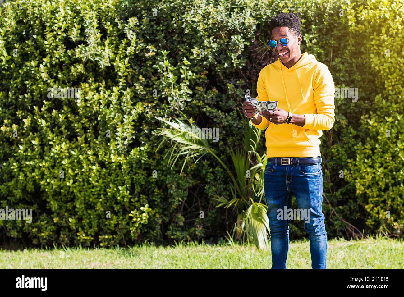 Ein lächelnder junger afrikanischer Mann, gekleidet in ein gelbes Sweatshirt, blaue Jeans und Sonnenbrille, der auf der Straße Geld zählt und Rechnungen zwischen seinen Händen übergibt. Stockfoto
