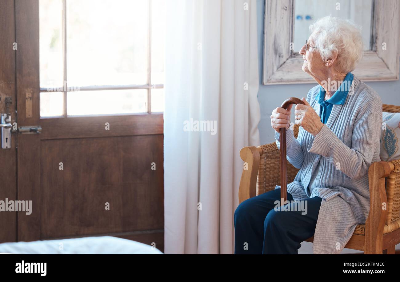 Denkende, traurige und ältere Frau, die an das Leben mit einem Spazierstock am Fenster im Zimmer eines Altershauses denkt. Ältere Frauen mit Depressionen Stockfoto