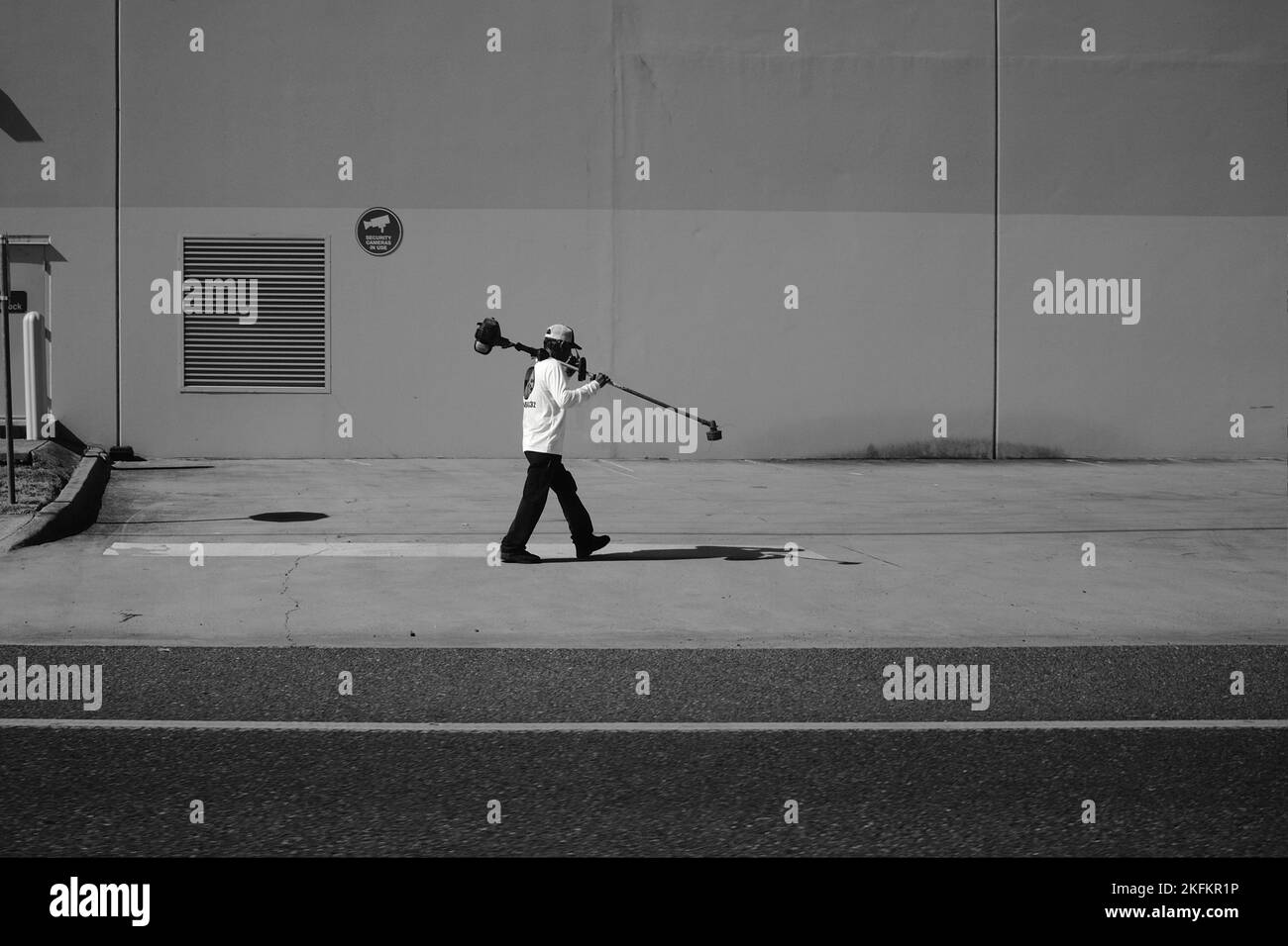 Eine Graustufe eines Gärtners, der vor einem Gebäude läuft und einen Unkrautfresser auf dem Schulterwandhintergrund trägt Stockfoto