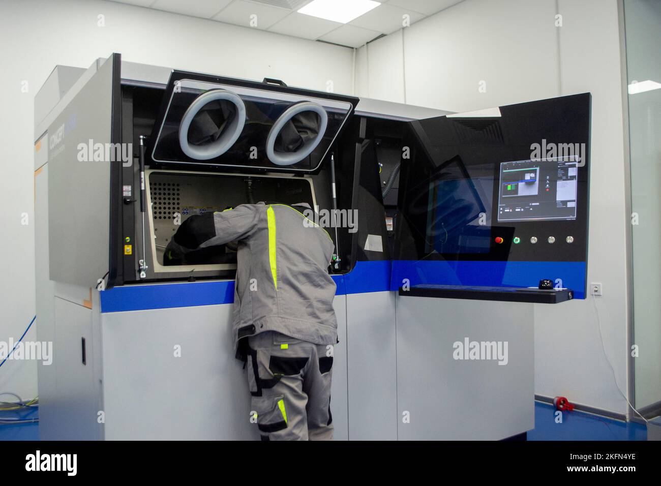 Ein Arbeiter blickt in ein offenes Fenster eines großen industriellen 3D-Druckers für Metallpulver. Der Arbeitsprozess. Industrial Powder 3D Drucker für Metall und Arbeiter Mann. Laser-Sintermaschine für Metall. Stockfoto