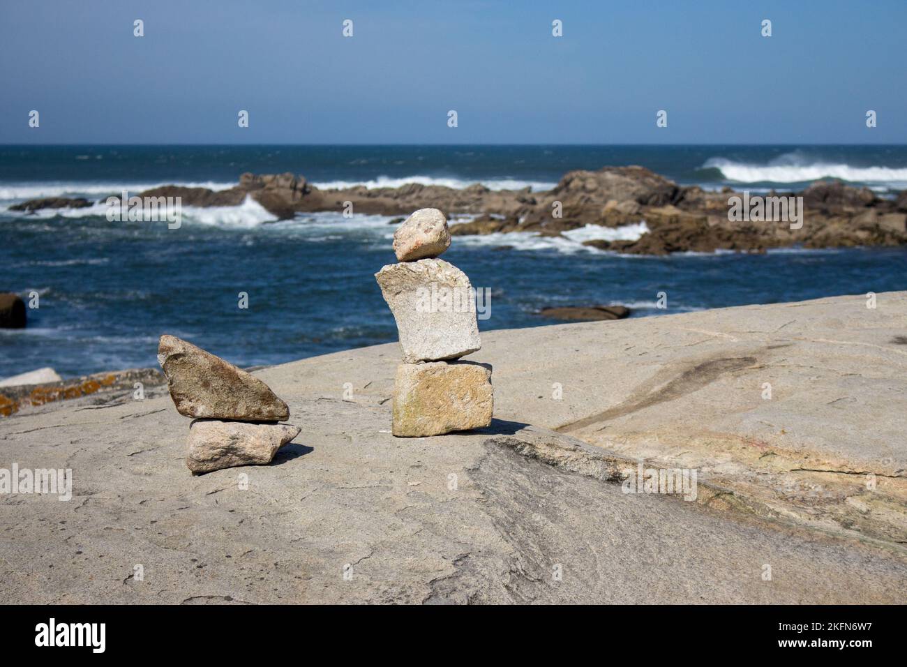 Turm aus kleinen Steinen an der Meeresküste mit Felsen im Hintergrund. Meereslandschaft mit Steinkunst. Konzept von Balance und Harmonie. Felsiger Strand mit blauen Wellen. Stockfoto