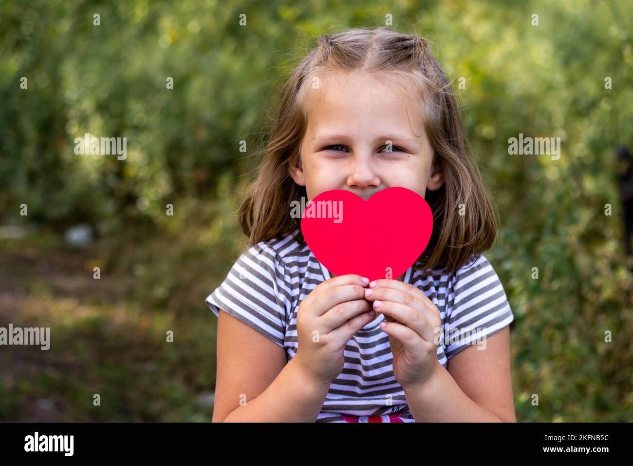 Ein Kind mit einem Papierherz in den Händen. Herzlichen Glückwunsch zum Valentinstag. Speicherplatz kopieren. Porträt eines süßen kleinen Mädchens im Park draußen. Liebe Stockfoto