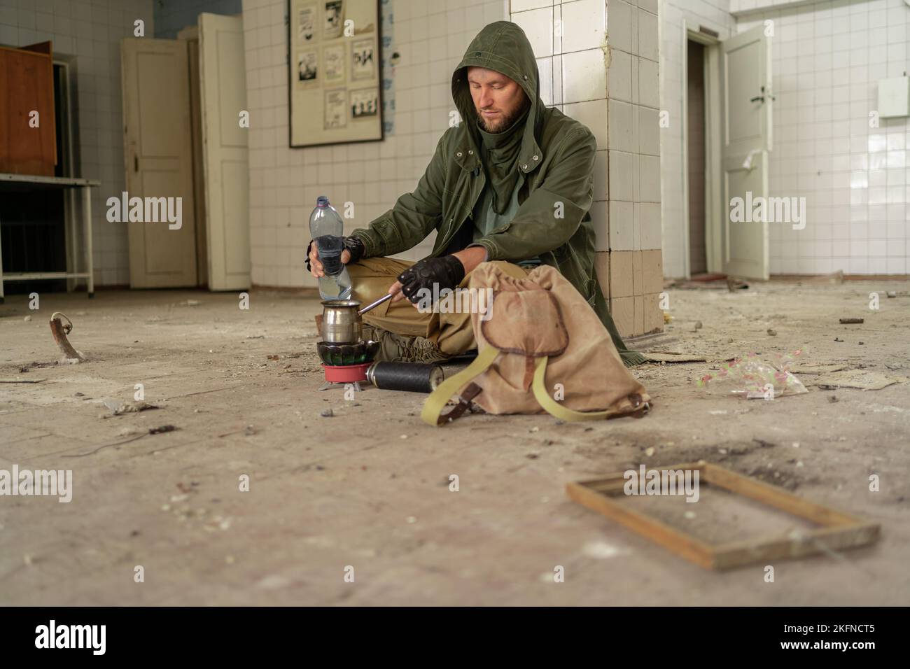 Post-Apokalypse. Ein junger Mann sitzt auf dem Boden eines verlassenen Gebäudes und gießt schweres Wasser in einen Becher. Konzept eines Mannes, der in einer post-apokalyptischen Welt wandert lo Stockfoto