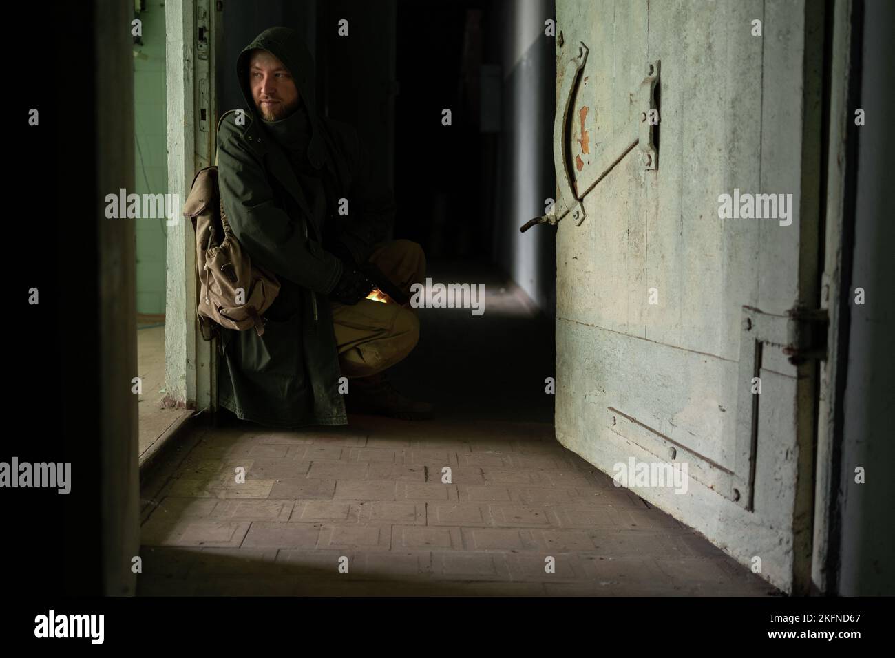 Ein schmutziger Mann in einer Kapuzenjacke sitzt neben der Tür in einem großen dunklen Korridor, hält eine Waffe in den Händen und schaut in den Raum. Das Konzept eines Überlebenden A Stockfoto