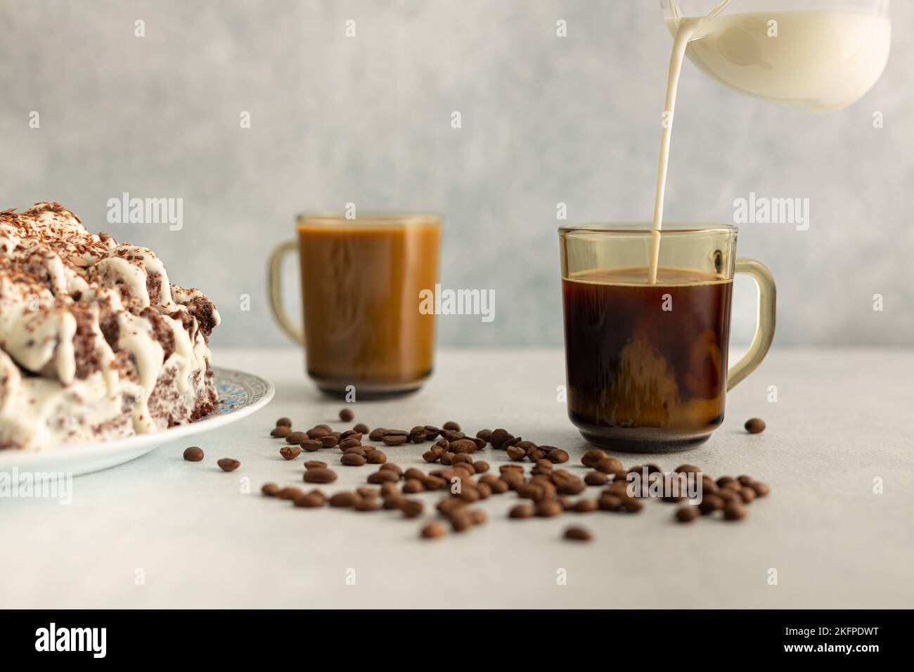 Kaffee mit Creme in einem Glas Becher mit Kaffeebohnen, Cremekuchen. Creme wird aus einem Glaskrug gegossen. Trinken Sie auf hellgrauem Hintergrund. Stockfoto