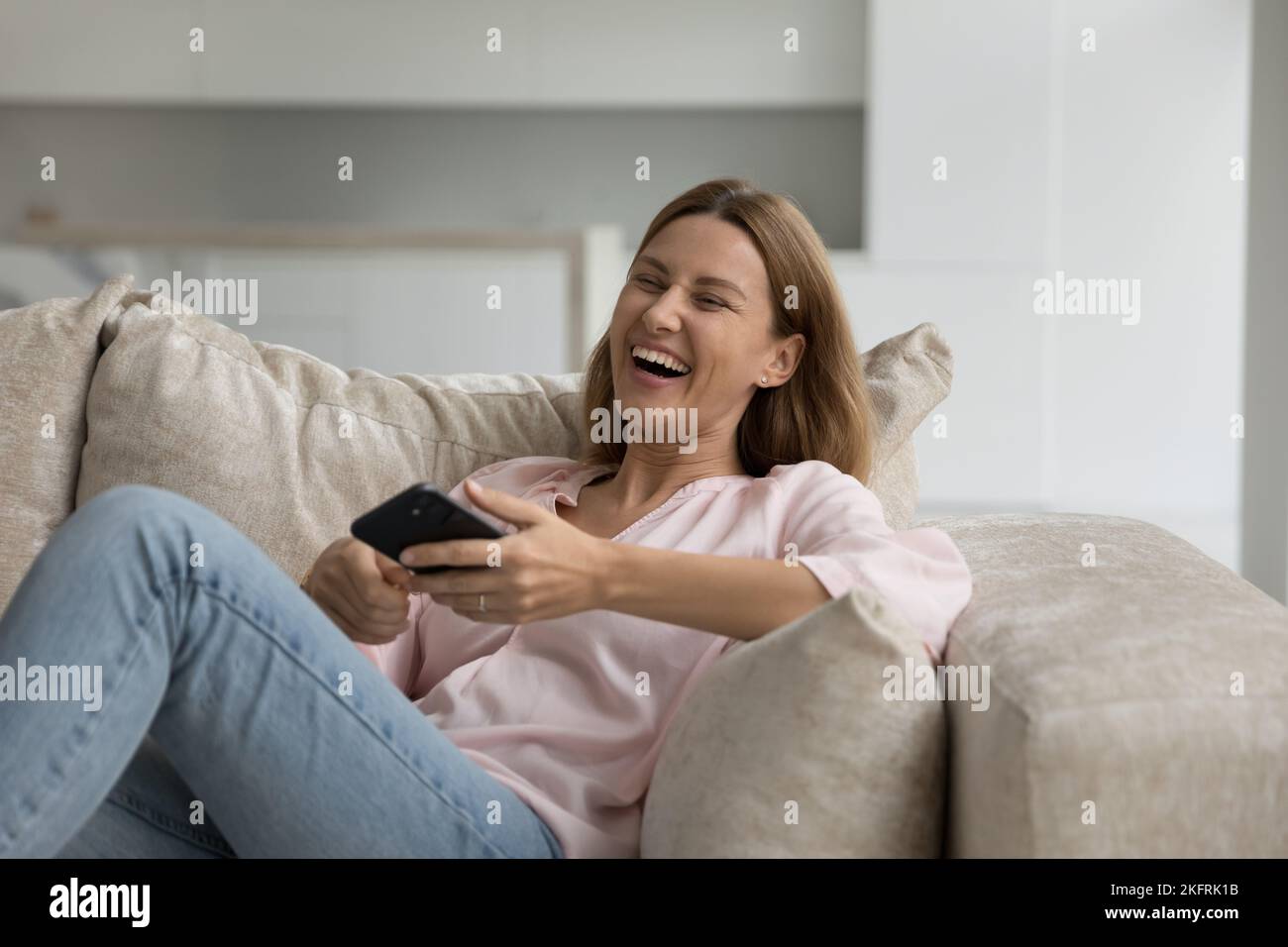 Fröhliche lachende Frau sitzt auf dem Sofa mit dem Smartphone Stockfoto