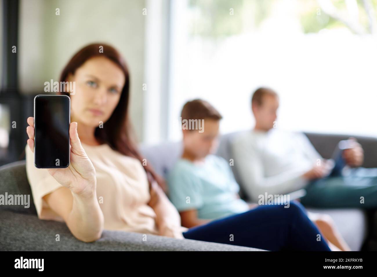 Zu viel Technologie... eine Frau hält ein Smartphone hoch, während ihre Familie mit ihren eigenen Geräten beschäftigt ist. Stockfoto
